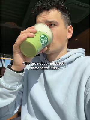 A man with short dark hair sips from a green beverage in a Starbucks cup, wearing a light blue hoodie. The background is blurred, with a hint of a sunny day.
