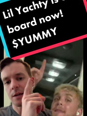 A selfie-style video cover with overlaid text. The image is divided into two sections. The top portion is dark with white text that reads, "Lil Yachty is on board now! $YUMMY" in bold, sans-serif font. The bottom section shows a man in the foreground, partially visible, pointing upwards with one hand, looking directly at the camera, and smiling. To the right of him is another person, also looking towards the camera and smiling. The background appears to be part of a vehicle interior. The overall tone of the image is promotional and enthusiastic.