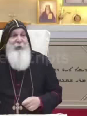 The image features an older man with a white beard, wearing a black religious garment. He is speaking directly towards the camera, with an ornate cross necklace visible. Behind him, there is a wall with what appears to be painted inscriptions with a painting of a religious figure hanging on the wall.