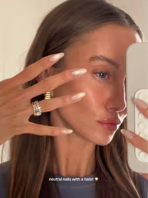 A close-up shot features a fair-skinned woman holding a phone in her hand while showing off her manicured nails. Her face is angled slightly towards the right, with her hand and nails in the foreground. The nails are a light color, reflecting the light. The image is well-lit, with a soft focus on the woman's face and a clear display of her nails. The background is blurred.
