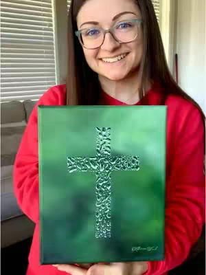 A woman with glasses smiles and holds a painting of a cross made of water droplets. She wears a red shirt. The background shows a window with blinds.