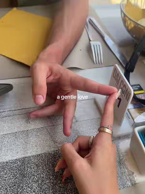 An overhead shot of two hands, one male and one female, almost touching. A male hand extends with a finger touching the female's finger, forming a heart shape. They are at a table in a restaurant setting, details like a menu and cutlery are visible. A text overlay reads 'a gentle love'.
