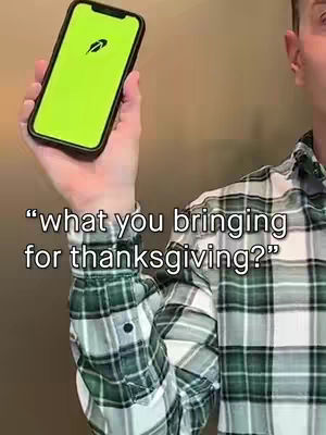 A man in a green and white plaid shirt holds a smartphone with a bright green screen, text overlayed on the image asks a question about Thanksgiving. The background appears to be a simple wall.