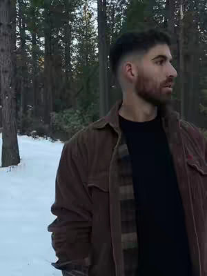 A man stands in a snowy landscape, his gaze turned towards the right. He is wearing a brown corduroy jacket over a black shirt and a plaid shirt underneath. The background features a forest with tall trees against a bright, overcast sky.