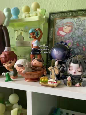 A variety of figurines are arranged on a shelf, including anime-style figures and other collectibles. The background appears to be a green painted wall, adding a pop of color to the setting. On the top shelf are cartoonish figures, with an artwork on display behind.