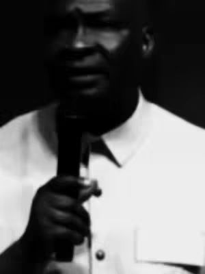 A grayscale image features a man, likely of African descent, holding a microphone, suggesting a public speaking event or a recording session. The lighting is dim, and the image is focused on the man's upper body, highlighting his face and the microphone, implying a focus on his words or message.