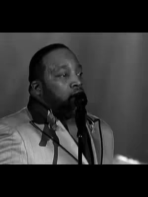 A black and white image featuring a man in a suit, holding a microphone. The background is blurred, indicating a performance setting.