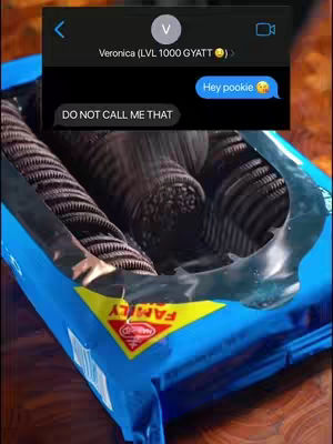 The image is a close-up view, capturing a package of Oreo cookies with a text message overlay. The perspective looks down into the open blue package with rows of dark chocolate cookies. The overlay consists of a black box representing a phone's text message interface, with a blue chat bubble containing the text 'Hey pookie' with a smiling face emoji, and a darker message bubble that says 'DO NOT CALL ME THAT'. Above the conversation, the phone's header displays a name 'Veronica (LVL 1000 GYATT)>' in white text, the text elements are set against a black backdrop, creating a contrast with the cookies and packaging.