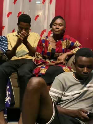 Three people sitting on a couch. The person in the middle are wearing a dress with red, blue, and yellow patterns, and the people on the either side are holding their hands up.