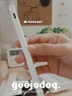 A hand holding a white stylus pen is the central focus, set against a softly lit background. Below the hand is a blurred display of items, including a digital clock, and what appears to be packaging. The top portion of the image is a backdrop with a Snoopy illustration and leaves, setting a clean, light aesthetic.