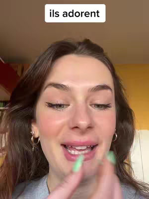 The cover image is a close-up shot of a woman's face, positioned with the camera angled slightly downward. Her skin appears to be smooth, with a light complexion. The woman has dark brown hair. She is wearing makeup, including defined eyelashes, a subtle blush, and lipstick. She has gold hoop earrings. A text overlay reading "ils adorent" (they love) sits in the upper-center part of the frame, within a white rectangle. The background is blurred, with warm tones of orange and brown, implying a cozy indoor setting. The woman is smiling gently, looking directly at the camera with a friendly expression. Her lips are moving as if she's speaking.