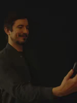 A man in a black shirt is holding a smartphone and looking at it while smiling. He is positioned on the left side of the frame with a dark background.