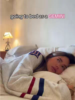 A woman lies in bed, looking thoughtful, wearing a gray sweatshirt with blue and red stripes on the sleeves. The text overlay reads, 'going to bed as a GEMINI'. A small lamp is visible in the background on her left.