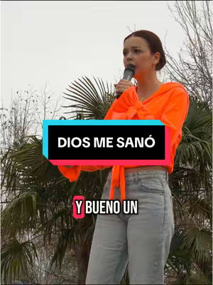 A woman in an orange top and light blue jeans is speaking into a microphone, positioned in front of a backdrop of green foliage and a cloudy sky. Text overlays announce the theme of faith.