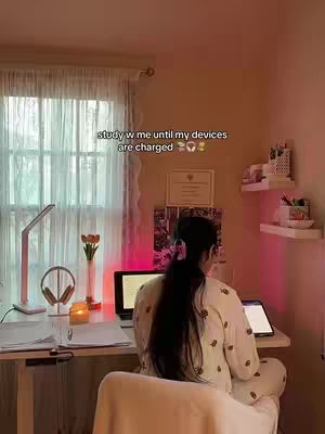 The image shows a woman from the back sitting at a desk, with a laptop and other study materials in a softly lit room. A window with sheer curtains fills the left side, and warm-toned lighting creates a cozy atmosphere.