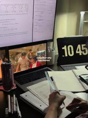 A well-lit desk setup with a laptop displaying a scene from a TV show, two computer monitors, a digital clock, several notebooks and pens, and a dark-colored can. The person appears to be studying, with hands visible, working while the show is on.