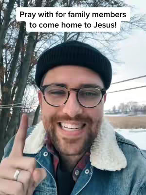 A man with glasses and a black beanie is smiling, giving a peace sign, and is gesturing towards the camera with a blurred background of trees and snow.