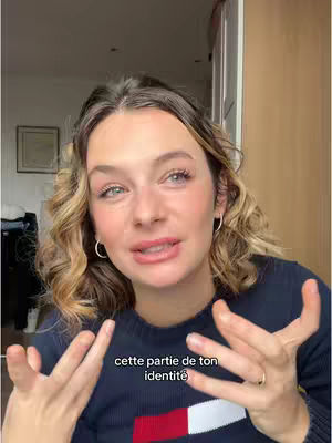 A woman with light skin and wavy blonde hair looks directly at the camera, speaking with an open and inviting expression, hands gesturing in front of her. She wears a navy blue sweater with a white and red emblem at the chest. In the background, a room is visible with objects like a picture frame and a door.