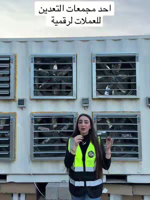 A woman stands in front of a white building, with multiple square windows featuring large fans that suggest a data center or crypto mining operation. She wears a high-visibility vest, suggesting a role related to maintenance or oversight. The image conveys a technological or industrial theme.