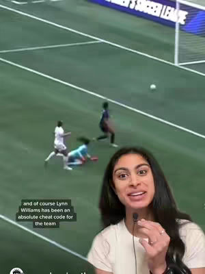 A woman with dark hair is filmed from the chest up, smiling while talking. A soccer game is displayed behind her, where players are visible and the goal net is in the background. Text appears in a box in the bottom left.