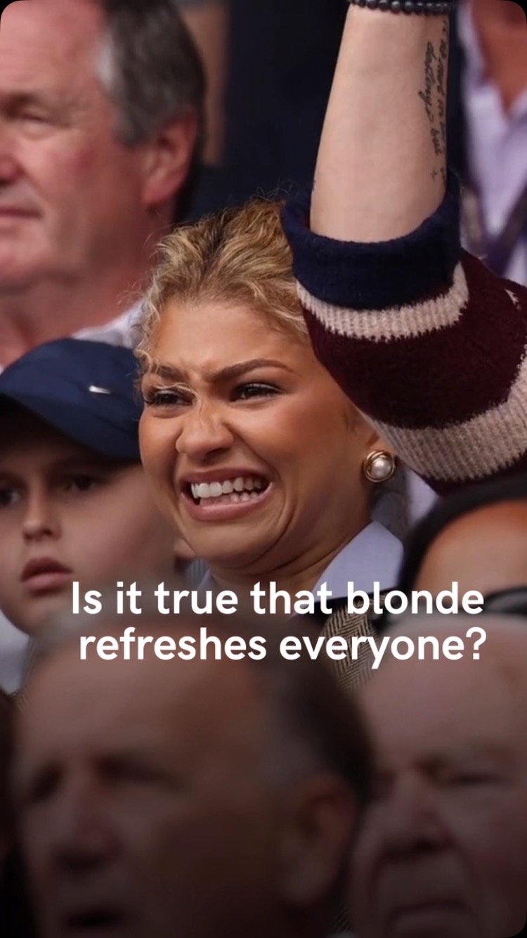 The image shows a woman, presumably with blonde hair, making a surprised or shocked facial expression. Her right arm is raised, and her upper arm with a sleeve and black beads is visible. The background shows a crowd of people watching an event.