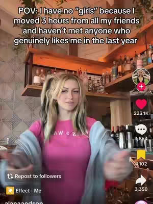 A young woman with blonde hair in pigtails stands in what appears to be a bar or restaurant setting. She's wearing a pink t-shirt under a light blue jacket, and she has a mildly expressive face and is gesturing outwards with both hands as if explaining something.