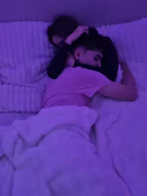 A couple is embracing in bed, under dim purple lighting. The man is lying on his back, wearing a pink t-shirt and covered partially by a white blanket. The woman appears to be hugging him from behind. The scene is intimate and bathed in purple light.
