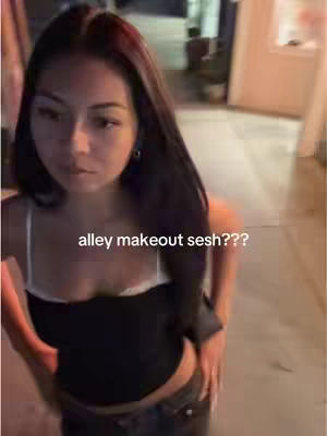 A young woman stands in an alleyway. The background is a blurred mix of light and shadow. The woman, who appears to be the main subject, is wearing a black crop top over a white spaghetti strap top and dark pants. The image is vertically oriented, and the composition is centered on her, with text overlaid at the center bottom.