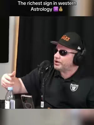 A man with a black cap and sunglasses is speaking into a microphone. The image has a podcast-like setting, with a microphone, a water bottle, and a phone visible. The background is dark.