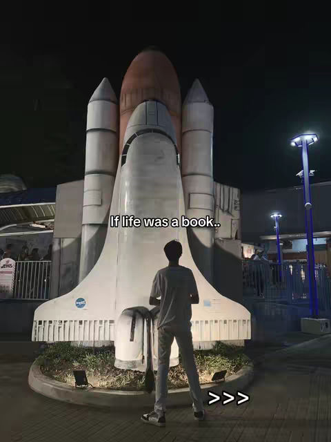 A man stands in front of a large space shuttle model at night. The shuttle stands with a neutral color palette, and the man has his back to the viewer.