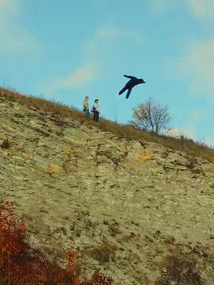 The image captures a landscape scene with a focus on a hillside and the sky. The hillside is primarily composed of a rocky, textured surface, dominating the lower portion of the frame. Two figures are standing on the hillside. In the upper right, a silhouetted figure appears to float in mid-air. The background consists of a clear blue sky. Soft, diffused light suggests an outdoor setting.
