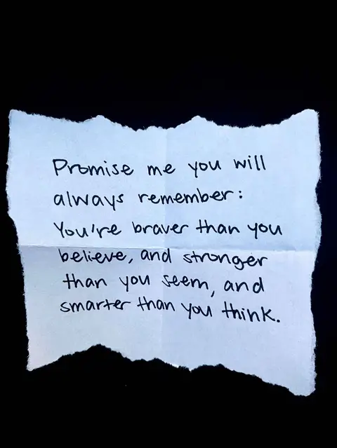 A close-up shot shows a piece of torn paper. The paper, slightly off-white, has handwritten text in black ink. The paper rests against a solid black background, emphasizing the text. The text includes the lines: 'Promise me you will always remember: You're braver than you believe, and stronger than you seem, and smarter than you think.'