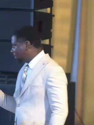 A man in a light-colored suit is standing and speaking, with his head turned to the side. The background consists of vertical structures and a yellow-orange gradient on the right.