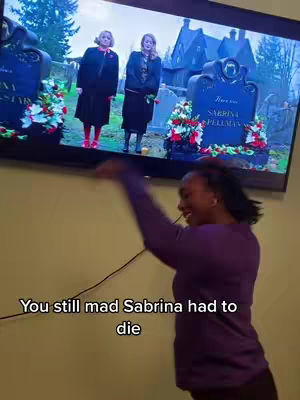A person standing in front of a TV screen showing a scene from the show Sabrina the Teenage Witch, where two women are standing in a graveyard. The person is gesturing with their arms and is wearing a purple shirt. Below the person, there is white text on the image.