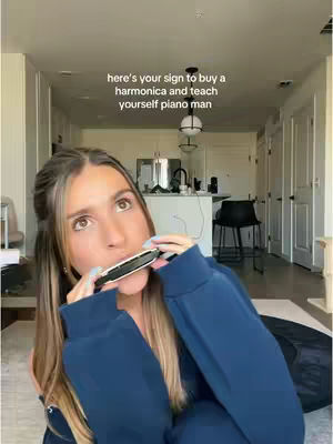 A woman is seated and playing a harmonica. She is wearing a dark blue hooded sweatshirt. Text overlay at the top of the image reads, 'here's your sign to buy a harmonica and teach yourself piano man'. The background shows a modern apartment with white walls, a kitchen, and a black chair.