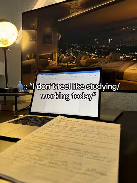 A cozy indoor scene is captured. A laptop displays a white screen with a text overlay and is center stage. A piece of paper with handwritten notes sits below the laptop keyboard. The background is a dimly lit room dominated by a large TV. A lamp on the side adds a spot of brightness to the scene.