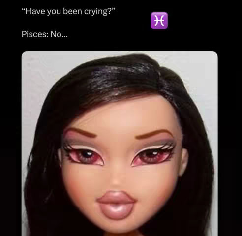 A close-up of a Bratz doll's face from the chest up against a black background. The doll has dark hair, full lips, pronounced eye makeup with red-toned eyelids. The eyelashes are long made-up.
