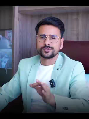 A man with glasses and a beard is speaking into the camera. He is wearing a light green blazer over a white shirt. Background appears to be wood paneling.
