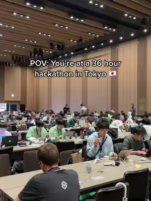 The image shows a large conference hall filled with people seated at tables, presumably working on laptops. The lighting is a mix of overhead lights and natural light from windows. The text overlay reads 'POV: You're at a 36 hour hackathon in Tokyo'.