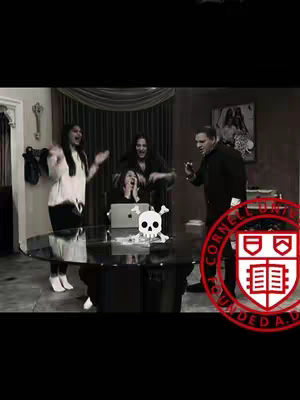The image is a black and white shot, with five people in a room. A round table is in the front with a skull placed on it. The main focus is on a group of people celebrating near a table with a laptop and a skull. A college logo is in the bottom right corner.