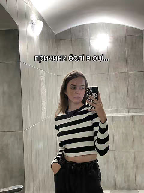 A mirror selfie shows a young woman in a bathroom. She is wearing a black and white striped long-sleeved shirt and black pants. She is holding a phone up to the mirror, taking the picture. The bathroom has plain, light-colored tile walls. Above the woman's head, text on the image reads 'prichiny boli v och...' translated to english as 'causes of pain in the eye...'