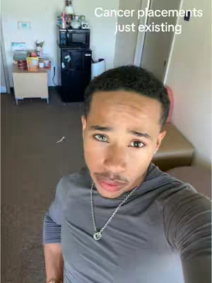 A selfie-style shot featuring a young Black man looking directly at the camera. The background is a casual indoor setting with domestic items and a microwave.