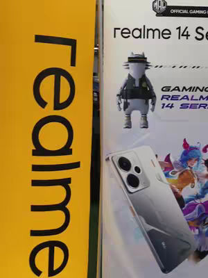 The cover photo shows a promotional display for Realme phones, set up in a retail environment. On the left, a vertical yellow banner displays "realme" in large, bold, black letters. To the right of the banner, the display features information about the Realme 14 series, including a cartoon figure and the phone model itself. The color scheme is bright, with yellow and white predominating.