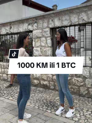 Two young women are standing on a cobblestone street, facing each other and talking. They are near a stonework building. A text overlay on a white rectangle says "1000 KM ili 1 BTC". One woman is wearing a white top, jean shorts, white shoes. The other is wearing a light tan top, jean shorts, and white shoes. The TikTok logo is visible on the left.