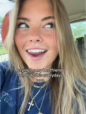 A close-up of a young woman looking to the side with open eyes and a wide smile. Her blonde hair is parted to the side and cascades down her shoulders. She wears multiple layered gold necklaces and a dark blue top. The background is slightly blurred, indicating the interior of a vehicle with a glimpse of greenery outside.