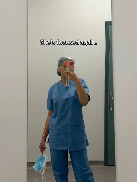 A mirror selfie shows a person wearing blue scrubs and a hair net, holding a smartphone in front of their face and a face mask. The background is a plain white wall and the reflection in the mirror. Text overlay says, "She's focused again."