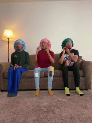 Three young men are seated on a brown couch in an interior setting. The lighting is soft, coming from a lamp on the left, illuminating the subjects and the background. The primary color scheme involves the skin tones of the individuals and the muted tones of the wall, rug, and couch. The individuals are wearing colorful hair coverings that stand out against their darker skin.
