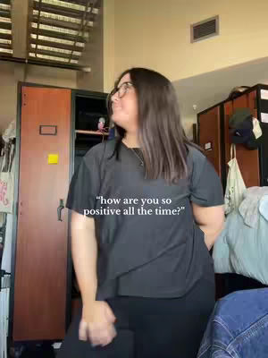 A woman wearing glasses and a black top is looking away from the camera against a backdrop of an apartment or dorm room. Text overlaid on the image reads, "how are you so positive all the time?".