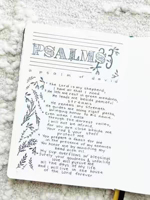 An open journal page is shown, featuring elegant calligraphy and floral decorations. The title 'PSALMS 23' is prominently displayed with decorative elements. The text of Psalm 23 is handwritten below, with stylized leaves and vines along the left side.