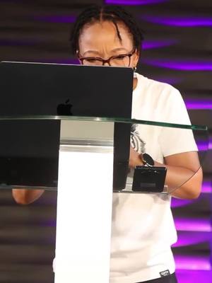 A woman with short, dark hair and glasses stands behind a podium, looking down at a laptop. She wears a white t-shirt and a watch. The background is a blurred purple and white.